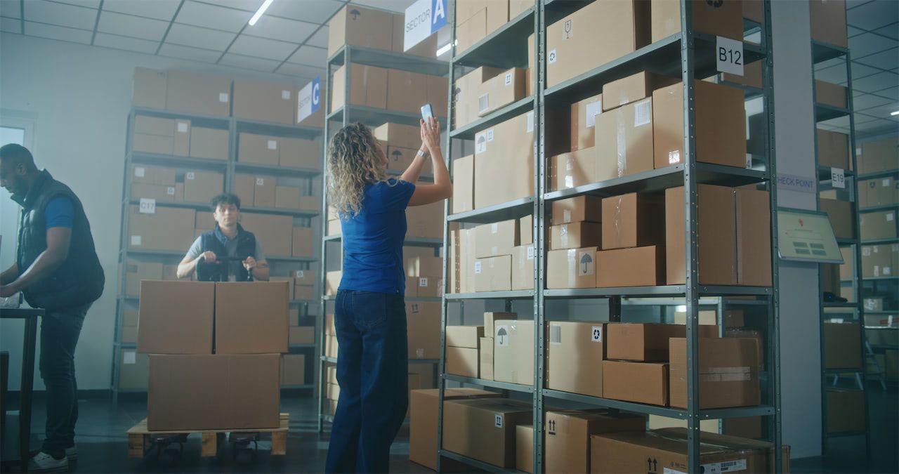 A woman takes a photo of stacked boxes on shelves in a warehouse, while two workers handle more boxes nearby.