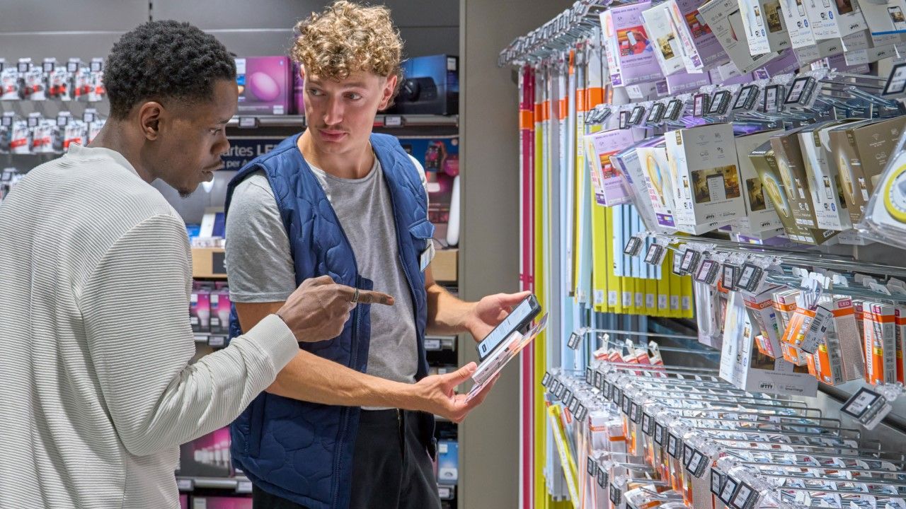 A customer in a white sweater points to a tablet held by a store employee in a blue vest, as they discuss products in an electronics aisle.