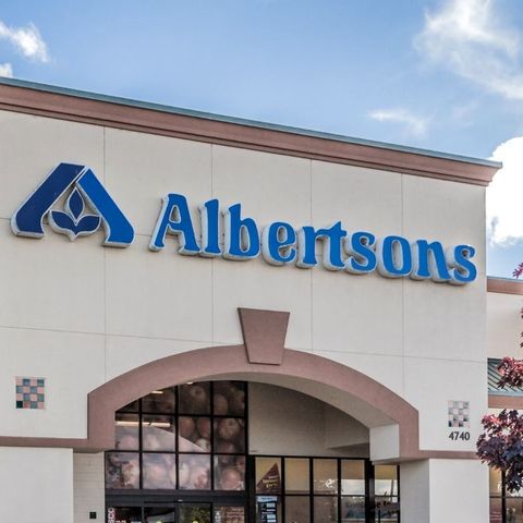 Front view of an Albertsons store with a prominent logo above the entrance. The building is surrounded by a clear sky and a tree.
