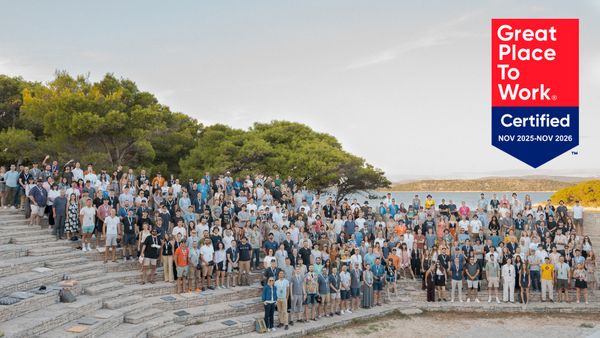 A large group of people stands on outdoor stone steps surrounded by trees and a body of water, next to a "Great Place to Work Certified" banner.