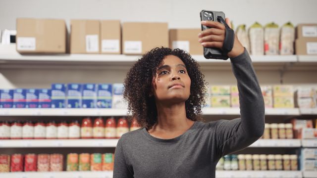 Woman in a grocery store aisle uses her smartphone, surrounded by shelves stocked with various products and boxes.