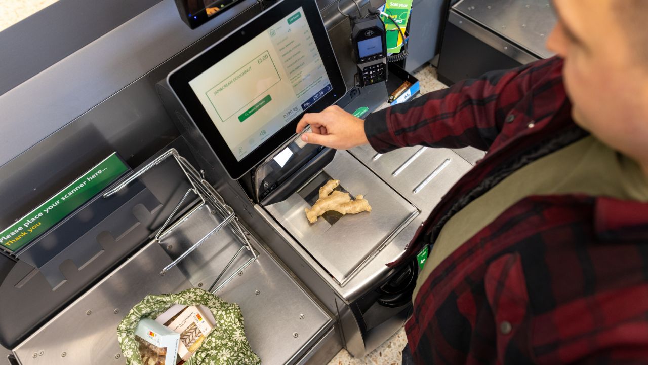 Person using a supermarket self-checkout kiosk, weighing fresh ginger root on the scale while touching the touchscreen display.