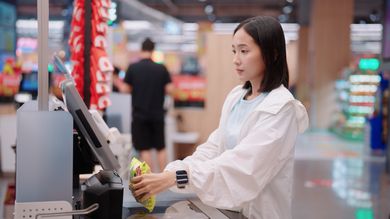 Young Asian woman in a white jacket using a self-checkout kiosk at a retail supermarket, scanning a food item with a smartwatch on her wrist.