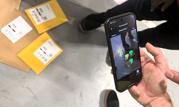 Person using a smartphone app to scan packages on the floor; three yellow envelopes visible nearby.