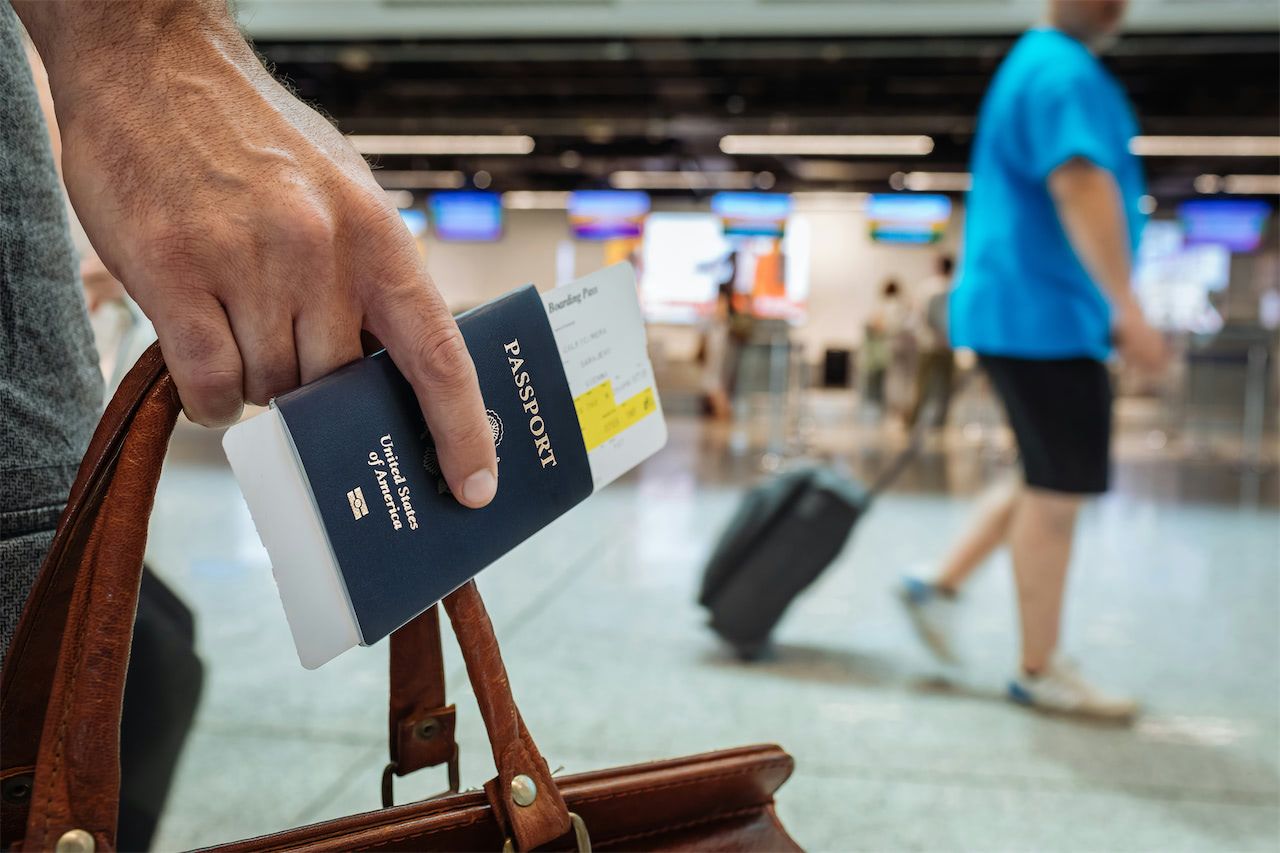 Close-up of a person holding a passport and boarding pass at an airport, with travelers and check-in counters in the background.