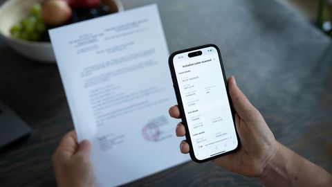 A person holds a smartphone displaying a vaccination certificate while holding a document. A bowl of fruit is in the background.
