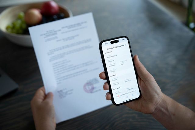 A person holds a smartphone displaying a vaccination certificate while holding a document. A bowl of fruit is in the background.