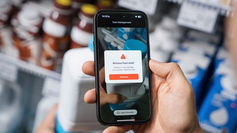 Person holding a smartphone displaying a "Remove from shelf" alert for a product, with shelves of items blurred in the background.