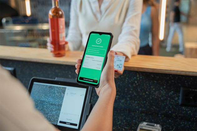 Cashier holding a smartphone displaying a green age and identity verification screen at a bar counter, with a customer holding an ID card in the background.