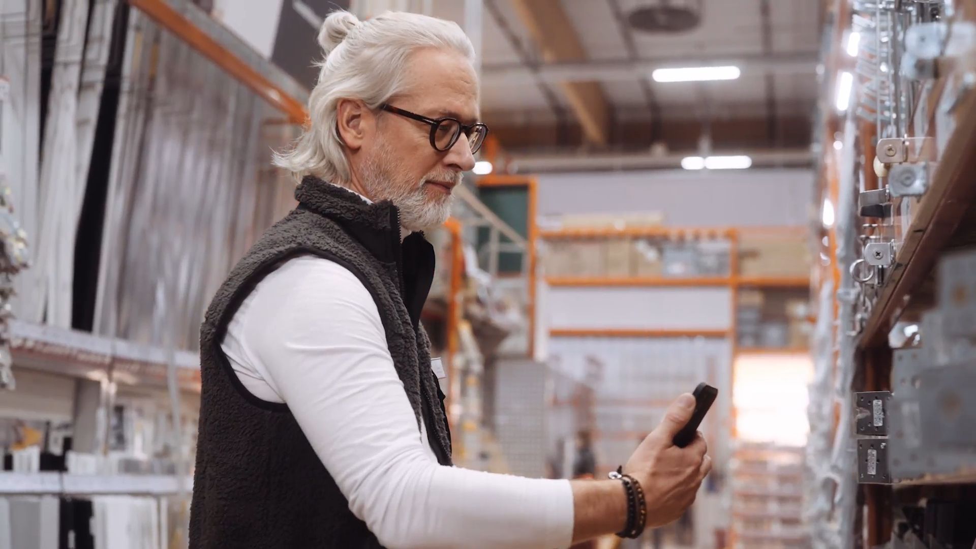 A man with grey hair tied in a bun and glasses uses a smartphone to check stock on tall shelves in a warehouse aisle.