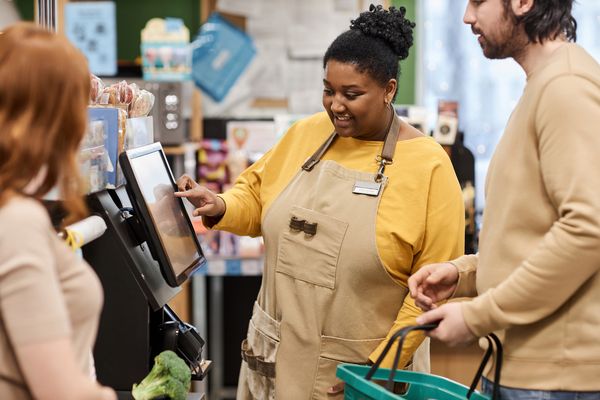 Smiling female grocery store cashier in apron assisting customers at a retail self-checkout register in a supermarket.