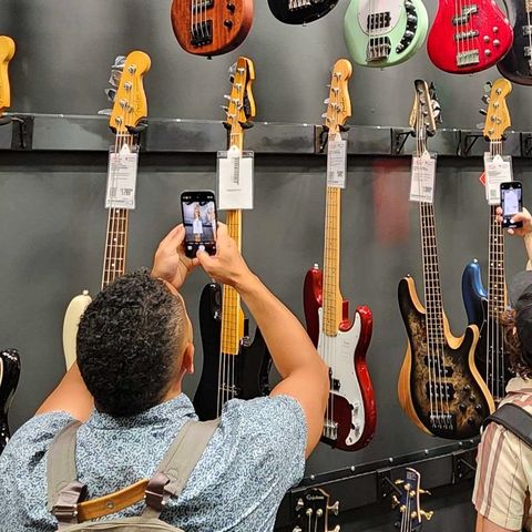 Two people take photos of electric bass guitars displayed on a wall in a music store.