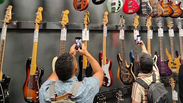 Two people take photos of electric bass guitars displayed on a wall in a music store.